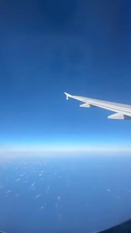 This media file features a view from an airplane window. The primary subjects are the airplane's wing, which is prominently visible extending across the upper right portion of the frame, and the vast expanse of the sky and clouds below.

The scene depicts a flight at a high altitude. The sky is a clear, deep blue, suggesting daylight. Below, a layer of white, fluffy clouds is spread out like a blanket over a deep blue ocean or sea. The horizon line is sharp and distinct, separating the sky from the cloud layer.

The time of day appears to be daytime, with the bright blue sky and visible clouds indicating good weather conditions. There are no discernible location cues beyond the aerial perspective, though the provided context specifies Yehud-Monosson, Israel, which is not visually evident in the image itself. The image evokes a sense of travel, journey, and possibly tranquility or awe associated with flying.

There is no visible text in the media file.
