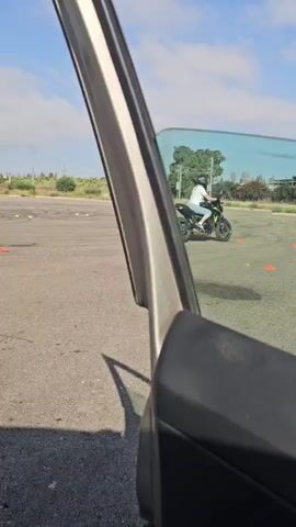 A person is riding a motorcycle in a parking lot or open area. The rider is wearing a helmet and a white shirt. There are orange cones set up in a pattern on the ground, suggesting a training course or obstacle course. The motorcycle is green and black. The sun is shining, and there are clouds in the blue sky. The scene is viewed from the perspective of someone inside a car, with the open car door and window visible in the foreground. The ground is asphalt, and there are some patches of dark stain. In the background, there are buildings with red roofs and trees.