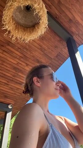 A woman wearing sunglasses and a striped swimsuit is looking up and adjusting her sunglasses. She is outdoors under a wooden roof structure with a thatched light fixture above. The sky is blue and there is greenery visible in the background. A security camera is mounted on a post to the left.