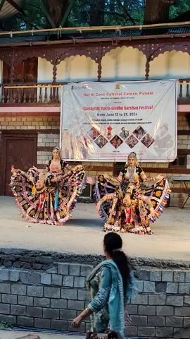 The video shows two women performing a traditional dance on a raised stage. They are wearing elaborate, colorful costumes with wide, flared skirts that appear to be made of multiple layers of fabric. Their outfits include ornate jewelry, headdresses, and armbands. The dancers are in dynamic poses, with one on the left with arms extended and the other on the right bent at the knees with arms raised. Behind them, a large banner is displayed that reads "North Zone Cultural Centre, Patiala" and "Sanskriti Yatra-Sindhu Darshan Festival FROM JUNE 13 TO 29, 2026". The stage is made of concrete or stone and is surrounded by brick walls. In the foreground, partially obstructing the view of the stage, is a person dressed in a patterned shawl and trousers, walking away from the camera. The background shows a wooden structure with arched openings and lush green trees. The lighting suggests it is either dusk or an indoor performance with artificial lighting.