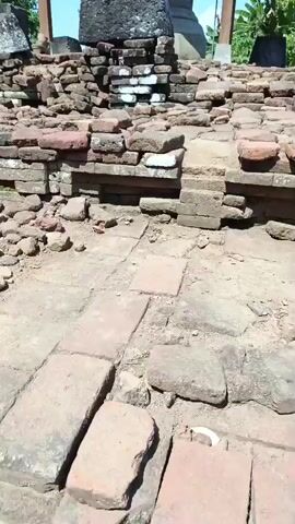The video pans across the ruins of an ancient stone structure. The foreground is dominated by scattered, weathered bricks and large stones, some of which form a crumbling pathway. In the midground, a larger, dark stone block, weathered and covered in lichen, stands atop a low wall of broken bricks. Behind this, partially sheltered by a wooden roof supported by pillars, is a large, weathered stone statue. The statue appears to be seated and is also covered in a patina of age. The background shows lush green trees and foliage under a clear blue sky. The overall impression is one of an archaeological site in a tropical or subtropical environment.