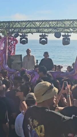 A crowd of people gather in front of a stage set up on a beach. The stage is decorated with purple and pink sculptures, and topped with a metal frame covered in greenery. Two DJs are at the front of the stage, playing music for the crowd. The people are dressed in a variety of casual clothing, and some are holding drinks or filming the event with their phones. Behind the stage is the ocean and a coastline. The sky is clear and bright, suggesting a sunny day.