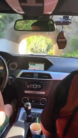 The video shows the interior of a car driving through a tunnel. The driver is holding a phone and recording. The passenger is looking at their phone. Sunlight can be seen at the end of the tunnel, illuminating trees and foliage. An air freshener hangs from the rearview mirror. The dashboard has a navigation screen displaying a map.