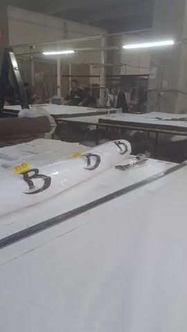 The video shows the interior of a large clothing factory, possibly in Turkey, with white tables for cutting fabric. There is a roll of fabric with "BD" printed on it, as well as scissors and tape on a table. Workers are seen standing at the tables, possibly cutting fabric. The lighting is fluorescent, and the environment is industrial. The camera is static and pans slightly.