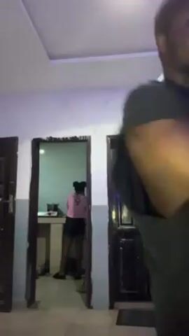 A young man in a dark-colored t-shirt and light-colored shorts dances in a hallway. He faces away from the camera, moving his arms to the music. There are three closed doors in the hallway. The door on the left and right sides are dark wood. The middle door is open, revealing a kitchen space with a young woman in a pink shirt and black shorts standing at the counter. A red towel hangs on the middle door. The room is painted light blue with a white ceiling. Artificial light is used.
