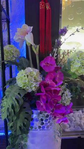 The video pans across a display of products and floral arrangements. On the left, a black metal shelf unit is visible, illuminated by blue LED lights. The shelves contain white boxes, some with text, and stacked items. One shelf holds a flyer with text and QR codes. In the foreground, a large floral arrangement features pink orchids, green hydrangeas, large green leaves, and a white vase with a mosaic pattern. Red decorative tassels hang from the top right of the frame. The wall behind the display has textured patterns and what appear to be Chinese characters. A magazine rack with several magazines is also visible on the left. The lighting is dim, with the blue LEDs creating a prominent atmosphere.