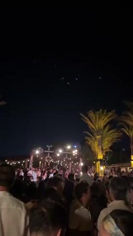The video shows a nighttime outdoor gathering of many people. The crowd is densely packed, suggesting a party or event. Palm trees are visible on the sides of the frame, and string lights and stage lighting illuminate the scene. The sky is dark, with some small, green lights appearing intermittently in the upper portion of the frame. The people in the foreground are looking forward, toward the illuminated area.