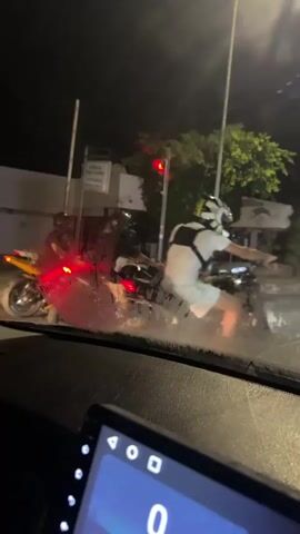 The video shows a nighttime scene from inside a vehicle, looking through the windshield. Two motorcycles are visible on a street. One motorcycle, which is yellow, is on the left side of the frame. Another motorcycle, which is dark colored, is to the right of the yellow one. A person is riding each motorcycle. The person on the dark motorcycle is wearing a white shirt and helmet. The person on the yellow motorcycle is wearing dark clothing and a helmet. Both motorcycles appear to be stationary or moving slowly at a traffic signal. The street is illuminated by streetlights and the headlights of the motorcycles. There are buildings and trees in the background. A traffic light is visible, showing a red signal. The windshield of the vehicle appears to be wet or dirty, causing some visual distortion.