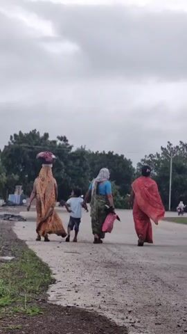 The video shows four people walking away from the camera down a dirt road. The person in the front left is carrying a basket of colorful items on their head. They are wearing a yellow and orange patterned dress. To their right, a young child in a blue shirt and dark shorts is walking. Next to the child is a woman in a green and black patterned dress, carrying a pink bag in her right hand. The person in the far right is wearing a bright red sari. The road is unpaved and has some gravel. To the left of the road is a patch of grass and some debris. In the background, there are trees and a cloudy sky. There is also a pole with wires on the right side of the road. The scene appears to be outdoors in a rural setting.