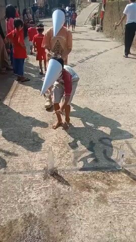 The video shows children participating in a game outdoors. They are wearing white, cone-shaped hats that obscure their vision. They are attempting to pour water from a cup into a plastic bottle on the ground. Other children and adults are watching. The ground is concrete.