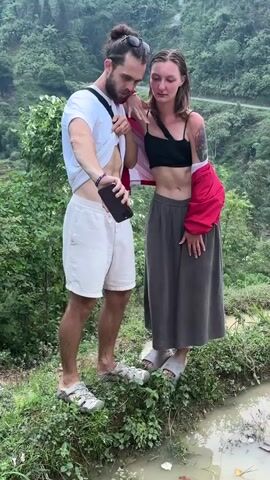 A man and a woman are taking a selfie outdoors in a lush, green, mountainous landscape, characteristic of Nậm Dịch Commune, Vietnam. The man, wearing a white t-shirt, white shorts, and sandals, holds a black smartphone with his right hand, pointing it towards himself and the woman. He has lifted his t-shirt to expose his abdomen. The woman, wearing a black crop top, dark wide-leg pants, and sandals, has a red jacket draped over her shoulders. Both subjects have pleasant, relaxed expressions, with slight smiles. They are standing on a narrow, grassy patch next to a small channel of water, possibly an irrigation ditch. Dense green vegetation, trees, and distant hills or mountains form the backdrop, suggesting a scenic, rural environment with potential for rice paddies.