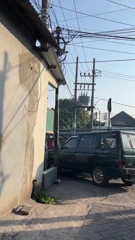 A dark green SUV is parked on a paved surface adjacent to a weathered concrete wall and a building with a corrugated metal roof. Overhead, a complex network of electrical wires and poles, including a transformer, dominates the sky. Sunlight casts shadows, indicating daytime. The car is positioned at an angle, partially obscured by the wall. There are no people visible in the frame. The environment appears to be an urban or semi-urban setting with signs of utility infrastructure and aging structures.