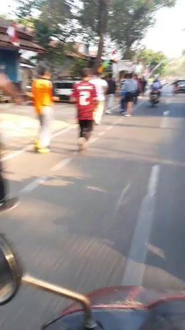 The video shows a street scene with several people walking and running down the road. There are also cars and motorcycles on the road, and flags are visible on the side of the road. The sun appears to be setting, casting long shadows. The perspective of the video suggests it is being filmed from the back of a motorcycle.