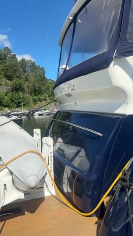 The video begins with a close-up of the stern of a white and navy blue yacht docked at a wooden pier. A yellow power cord is attached to the yacht and runs along the deck. The camera then pans to the side of the yacht, showing its hull and some portholes. In the next clip, a woman wearing a white t-shirt and black shorts walks away from the camera down the pier, carrying a tote bag. She walks past several docked boats, including sailboats and a motor yacht. The video ends with a view of sailboats and other vessels docked in a harbor under a clear blue sky with some scattered clouds. The water is calm, and trees and buildings are visible on the shore in the distance.