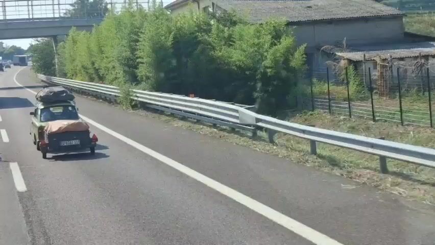 A green Trabant car is driving down a highway, pulling a small trailer. Both the car and the trailer are loaded with luggage, covered with tarps. The car is in the right lane, and a metal guardrail runs along the left side of the road. Trees and bushes line the other side of the guardrail. An overpass is visible in the background.