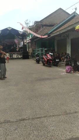 This media file captures a street scene in Kibin, Indonesia, likely during a daytime event. The image is oriented vertically. The main focus is a street lined with buildings, some appearing to be shops or homes with tiled roofs. A blue tent is set up in the street, indicating an organized event.

Under the tent, a stage is visible with sound equipment, suggesting a performance or gathering is taking place. Several people are present, though their specific actions are difficult to discern from this angle. In front of the stage, there are stacks of speakers and some lighting equipment.

Several motorcycles are parked along the street, both in front of and behind the tent. One prominent red motorcycle is parked near the center of the frame. A few individuals are seen standing or sitting near the motorcycles and in the background. One person in a red shirt is walking away from the tent area. The presence of motorcycles and the style of the buildings suggest a typical Indonesian street environment.

Decorations, possibly flags or banners, are strung across the street in front of the tent, adding a festive atmosphere. The sky appears overcast, suggesting it might be a cloudy day or potentially a break in the rain. The overall scene conveys a sense of community activity and local celebration. No legible text is visible in the frame.