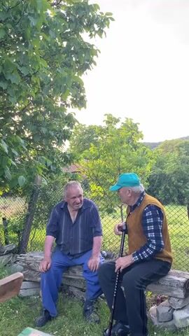 Two older men sit on a wooden bench outdoors, talking. The man on the left wears a blue striped shirt and blue pants, smiling at the other man. The man on the right wears a blue and white plaid shirt, a brown vest, and a green cap, holding a walking stick. They are surrounded by lush green foliage and a wire fence. In the background, rolling green hills under a partly cloudy sky are visible.