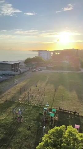 The video pans across a grassy field at sunset, showing a playground with several children on it, and a group of people playing soccer on a field further away. The sun is setting over the ocean in the background, casting long shadows across the scene. There are buildings and trees visible in the distance.