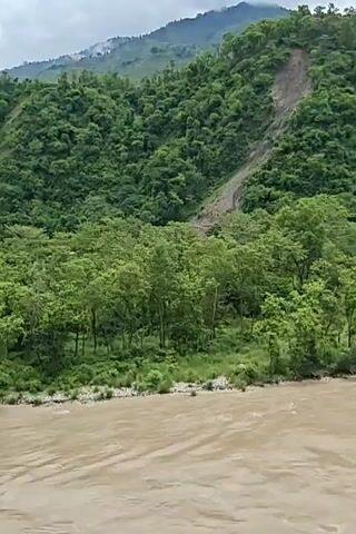 The video shows a wide, muddy river flowing through a lush, green valley. The riverbanks are lined with dense trees, and steep, tree-covered mountains rise in the background. The sky is cloudy, with some patches of blue visible. There are no people or man-made structures visible in the immediate foreground or river. A large section of the mountainside appears to have been affected by a landslide, with a scar of exposed earth and debris running down the green slope. The river appears to be swollen, possibly due to recent rainfall, and the water is flowing rapidly. The overall impression is one of natural beauty and the power of water, with a hint of the destructive force of nature evident in the landslide.