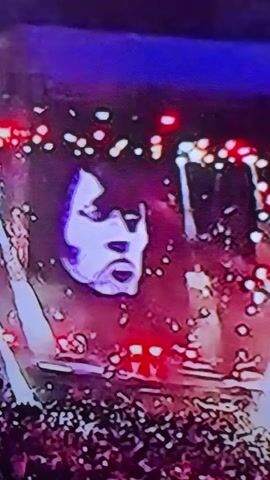 A digital projection of a stylized, white face with dark eye makeup and sharp features is displayed against a dark background illuminated by red and purple lights. The face appears to be that of a male with a shocked or screaming expression. Below the projection, a blurred, dark mass of people is visible, suggesting a concert or large event. The lighting is dim and atmospheric, with scattered bright lights resembling stage lighting or a crowd holding up lights.