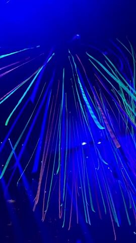 The video shows long, thin ribbons hanging from a dark ceiling, illuminated by neon blue and ultraviolet (UV) light. The ribbons appear in vibrant shades of neon green, blue, and pink/orange, creating a glowing, ethereal effect against the dark background. Blue light beams are also visible, cutting through the space and adding to the atmospheric lighting. Some light fixtures are faintly discernible among the ribbons. The scene suggests an indoor party or event, possibly a club or similar venue in Nir Eliyahu, Israel, given the themed lighting and the lively, ambient music playing in the background. No people or visible text are present in the frame.