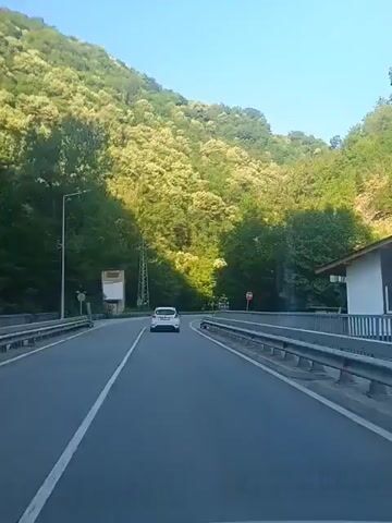 The video captures a first-person view from inside a moving vehicle. The vehicle is traveling on a paved road that curves to the right. The road is flanked by guardrails on both sides. The terrain to the left of the road rises steeply and is covered in dense green trees, with some trees bearing clusters of light-colored blossoms. A tall metal utility pole is visible on the left side of the road, near a large rectangular sign with a white background and orange trim. A smaller road sign with a white triangular shape is also present on the left. To the right of the road, the guardrail continues, and beyond it, the terrain is also covered in trees. A white car is visible ahead, also traveling on the same road. The sky is clear and blue.