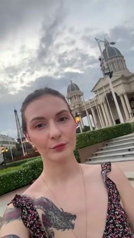 The video shows a selfie taken by a woman with brown hair and fair skin, wearing a floral dress and a dragon tattoo on her left shoulder. She is looking directly at the camera. Behind her, a grand building with classical architecture, columns, and domes is visible. The building appears to be a government or public institution. The woman is standing on a paved area with steps leading up to the building. The sky is cloudy, suggesting it might be late afternoon or early evening. The camera movement is slight, creating a subtle panning effect.