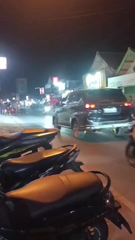 The video shows a nighttime street scene with several parked motorcycles in the foreground. In the background, there is traffic on a road with illuminated shops and buildings on either side. Several people are visible riding motorcycles and walking along the street. The lighting suggests it is dark, with artificial lights from the shops and vehicle headlights illuminating the area. The overall atmosphere appears to be that of a busy street at night.