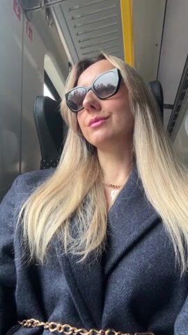A woman with long blonde hair and sunglasses sits in a train seat. She wears a navy blue coat and a gold necklace. She is applying pink lipstick with a small applicator. The train interior is visible in the background with a yellow handrail and metal ceiling panels.