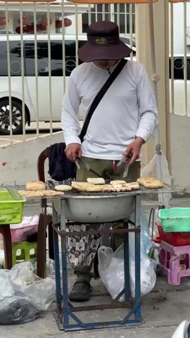 A person wearing a wide-brimmed hat, a long-sleeved white shirt, and olive green pants is grilling food outdoors. The food appears to be flat, oval-shaped items placed on a metal grate over a charcoal grill. The person is using tongs to turn the food. The grilling setup is on a street with a white fence and a white vehicle in the background. There are plastic crates and bags around the grilling area.