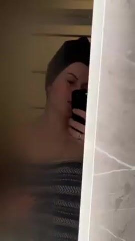 A woman is taking a selfie in a steamy bathroom mirror. She has her hair wrapped in a dark towel and is wearing a towel around her body. She is holding a black smartphone in her right hand. A wedding band is visible on her left ring finger. A hand reaches into the frame and wipes a portion of the mirror, improving visibility. The background shows a tiled wall.