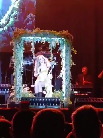 A woman wearing a yellow dress and black boots is singing into a microphone while sitting on a swing on a stage. The swing is enclosed in a square frame decorated with greenery and lights. A band is playing behind her. The audience can be seen in the foreground.  The lights from the crowd’s cellphones are on.