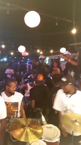 The video shows a crowded party at night with a live band. The scene is dimly lit with round white and orange lights hanging from the ceiling. Several people are dancing and standing around tables. A drummer is visible in the foreground, playing a drum kit. Several men wearing hats appear to be performers or party hosts, one holding a microphone.  Someone is exhaling a large cloud of smoke.  The atmosphere is lively and festive.