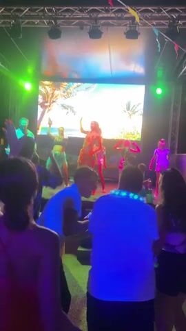 A group of people are dancing on a stage in front of a large screen displaying a tropical scene with palm trees and the ocean. The stage is lit with colorful lights, including green, pink, and blue. Some of the dancers are wearing brightly colored outfits. A crowd of people are watching from the audience, with some also dancing and holding up glow sticks. The atmosphere appears to be festive and energetic.