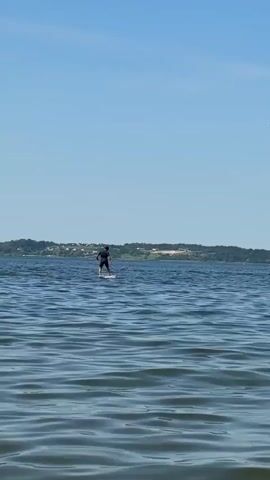 A person stands on a paddleboard in the middle of a body of water. The person is wearing dark clothing and appears to be paddling. The water is blue and calm with small waves. In the background, there is a treed shoreline with some buildings visible. The sky is clear and blue.