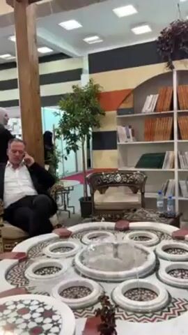 The camera is positioned in a room or booth, focusing on a central, ornate fountain. The fountain is circular with multiple raised, ring-shaped tiers. Water is flowing or bubbling in the fountain. Around the fountain are ornate chairs and a seating area. A man, dressed in a white shirt and dark jacket, is seated in one of the chairs and is talking on a cell phone. To the left of the man is a wooden post, possibly part of a structure. There is a small plant on a stand nearby. Behind the fountain, there is a bookshelf filled with books. The backdrop consists of striped black, white, and golden yellow panels. There is an opening that reveals a different area. The overall impression is that this might be a booth at a trade show or an exhibition.