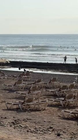 The video shows a beach with rows of empty lounge chairs in the foreground. In the middle ground, there is a dark, rocky shoreline with a small stream of water flowing into the ocean. Several people are in the water, some appearing to be surfing. Two individuals stand on the shoreline, facing the ocean. The ocean itself has moderate waves, and the sky is clear and bright, suggesting daytime. The overall scene depicts a beach resort area with recreational activities.