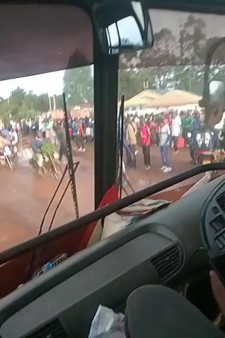 A camera in the front cab of a bus films a muddy road ahead filled with people, motorcycles and other vehicles. The crowd appears dense in the center of the frame, with people wearing a mix of light blue, red, and white clothing. A green pickup truck slowly moves through the crowd. The background includes trees, and buildings. The camera is stationary.