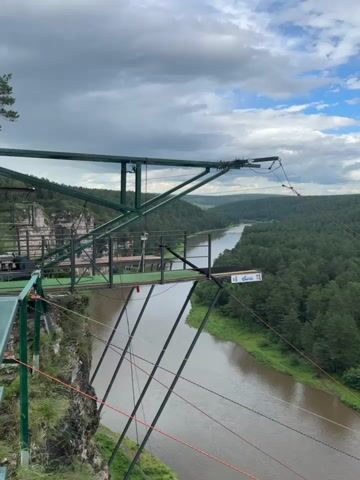 This media file captures a breathtaking view from a high platform overlooking a wide, winding river that flows through a dense, lush green forest. The platform, a robust metal structure painted green, extends out over a rocky cliff face. Cables, both thick and thin, are prominent, some extending horizontally from the platform, suggesting a zipline or bungee jumping setup.

The river, a murky brown, snakes through the valley, its banks lined with vibrant green foliage. The forest is a sea of tall, dark green pine trees, stretching as far as the eye can see and forming a dramatic backdrop. In the distance, rolling hills cloaked in the same dense forest fade into the horizon under a dramatic sky.

The sky itself is a dynamic mix of white, fluffy clouds and patches of bright blue, suggesting a partly cloudy day, possibly late afternoon or early evening given the diffused light. There are no people immediately visible on the platform or in the surrounding area, but the presence of the equipment strongly implies an activity like ziplining or bungee jumping is either about to happen, has just occurred, or is regularly offered here.

A small, white rectangular sign is attached to the edge of the platform, featuring a blue and white logo and some text that is difficult to decipher from this distance but appears to be an advertisement or informational sign for the attraction.

The overall impression is one of adventure and the raw beauty of nature. The height and the vastness of the landscape evoke a sense of awe and excitement. The setting, identified as Kul'metovo, Russia, points to a location offering outdoor recreation amidst stunning natural scenery. The lack of visible people, combined with the active zipline infrastructure, creates a feeling of anticipation.