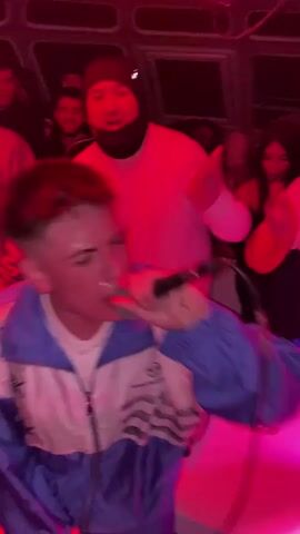 A young man is singing into a microphone in what appears to be a crowded nightclub or party venue. He is wearing a blue and white track jacket over a white t-shirt. He has short, light brown hair and some visible tattoos on his neck. Several other people are gathered around him, some dancing or moving to the music, while others are observing. One man, wearing a white sweater and a black beanie, is standing directly behind the singer. The lighting is primarily red, with some pink hues as well. There are water bottles visible on a table in the foreground. An "emergency exit" sign is visible on the ceiling.