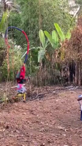 The video shows a person dressed in a colorful costume dancing in a grassy area with trees. The dancer wears a red top, blue shorts with a patterned skirt, and multicolored leg coverings. A tall, flexible headpiece with red, blue, and black ribbons arches above them. They carry a short stick in their right hand. Several men are seated on chairs watching the dancer, with at least one appearing to be drinking from a bottle. The setting appears to be in a rural or semi-rural location, with a low wall and trees in the background. There is trash scattered on the ground.