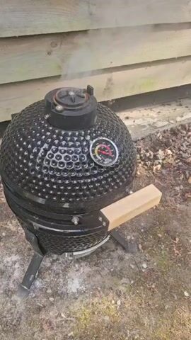 A black ceramic kamado grill is shown in an outdoor setting, with a wooden wall in the background. Smoke is rising from the top vent of the grill. The grill has a thermometer on its side, which appears to be indicating a temperature in the range of 200-250 degrees Fahrenheit. A wooden handle is attached to the side of the grill. The ground around the grill is covered in dirt and scattered leaves. The video pans slightly to the right and zooms in on the grill, focusing on the thermometer and the rising smoke.