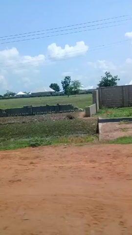 The video shows a landscape with a dirt road in the foreground. In the mid-ground, there is a broken concrete wall. Beyond the wall, there is a grassy field with scattered trees and several white-roofed buildings. The sky is blue with some clouds. Power lines are visible in the upper part of the frame. The video appears to be filmed from a moving vehicle, as indicated by the slight motion blur and the changing perspective.