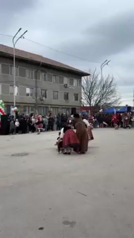 The video captures a traditional Bulgarian folk celebration taking place in a town square. There's a group of individuals dressed in folk costumes surrounding a small child seated on a chair. The adults move around the child while engaging in traditional dance steps. Behind them, a crowd of spectators watch the performance, and some figures are recognizable in festive costumes with tall, elaborate headgear. In the background stands a gray, multi-story building. The weather seems overcast. Balloons and flags are visible amongst the crowd. The overall impression is a festive, cultural event.