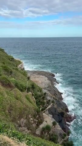 The video showcases a scenic view of a rocky coastline meeting the ocean under a partly cloudy sky. The camera slowly pans from left to right, revealing more of the vast blue-green sea.

In the foreground on the left, a steep, grassy cliff covered with lush green vegetation slopes down towards the water. Below the cliff, dark grey and reddish-brown rocks are visible, against which white foamy waves crash with considerable force, creating a dynamic contrast with the turquoise water. Scattered amidst the rocks and on the lower parts of the cliff are small, palm-like plants.

As the camera pans right, the expanse of the ocean dominates the frame. The water exhibits varying shades of blue and green, indicating depth and movement. Further in the distance, a prominent landmass, likely an island or a part of the mainland, is visible on the horizon, characterized by its rolling, green hills. The sky above is a mix of light blue with scattered white clouds. The overall scene depicts a rugged, natural coastal landscape with powerful ocean waves.