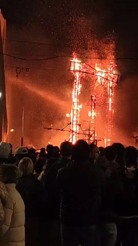 A crowd of people watches three wooden structures engulfed in flames against a dark night sky. Sparks rise from the burning structures. The crowd stands close, observing the spectacle. Power lines are visible above. The burning structures are tall and appear to be made of wooden planks.
