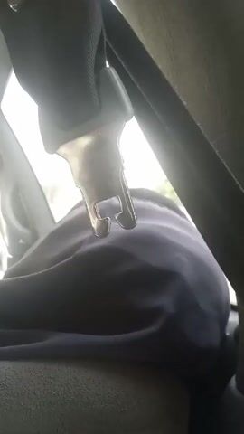 From an interior perspective of a vehicle, a seatbelt latch hangs down in front of a passenger, who is wearing a purple shirt. The camera is positioned close to the latch, capturing its metallic sheen and shape. The background reveals a view outside the car window, showcasing a road with other vehicles and buildings on the side. The lighting is bright, with sunlight filtering through the window.