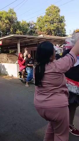 A woman in a pink tracksuit dances with another person dressed in a flamboyant costume. The costumed person has heavy makeup, a black top, and a multi-layered skirt of black and red material, with silver accents. They are holding what appear to be plastic bags or decorative elements. In the background, other people are present, some watching, and a red and white flag is visible on a pole. The scene takes place outdoors on a paved surface, with buildings and trees visible.