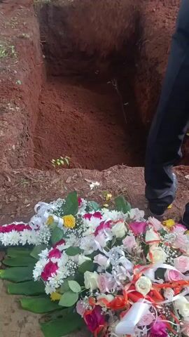 The video shows a freshly dug grave in red soil, with a large floral arrangement at the edge.  The arrangement includes roses, chrysanthemums, and greenery, decorated with ribbons. A person wearing black pants and brown shoes stands near the edge of the grave, partially obscuring the view of the hole.  There is some foliage and debris on the ground around the grave.