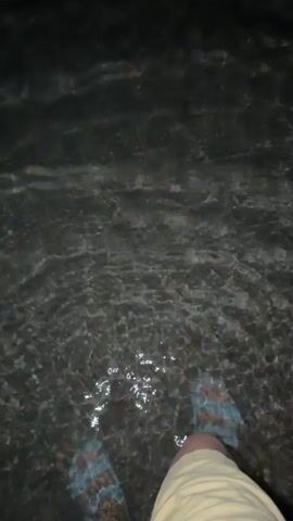This is a short video clip taken at night, likely at a beach or waterfront in Cholpon-Ata, Kyrgyzstan. The primary subject is the moonlit water.

In the first few seconds, a person's legs are visible as they stand in shallow water. They are wearing light blue sandals. The water is dark, with ripples catching the faint light. A small fish is briefly visible swimming in the water.

The scene then shifts to a wider view of the water. The moon is visible in the dark sky, appearing as a bright, luminous orb. Its reflection creates a shimmering, broken path of light across the surface of the water, extending towards the viewer. The overall mood is tranquil and serene, with the darkness of night contrasted by the bright moonlight on the water. There is no visible text in the video. The location context of Cholpon-Ata, Kyrgyzstan, suggests this could be a view of Issyk-Kul lake.